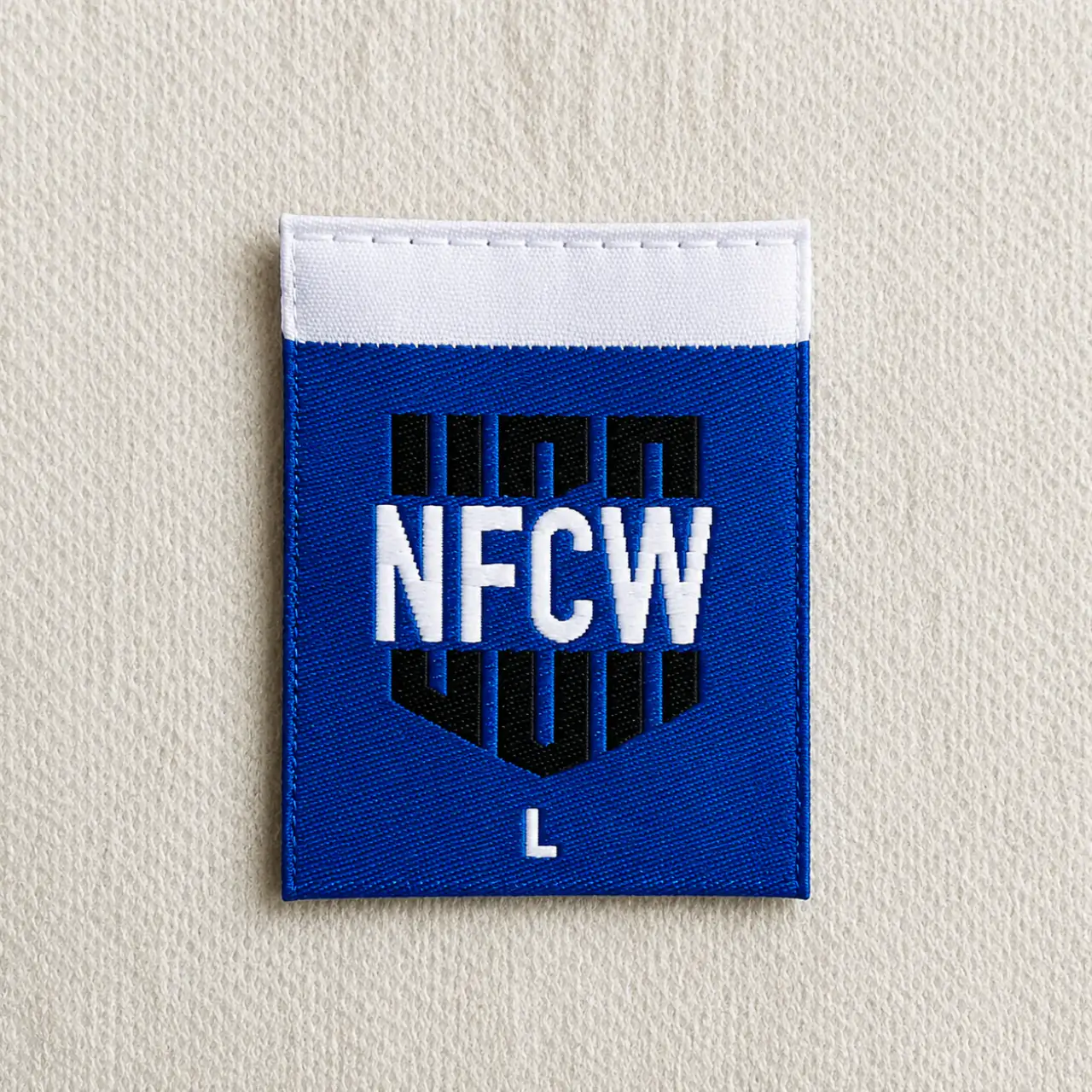 NFCW blue shield-shaped woven brand label with white wordmark, L size marker and white tab top — sportswear and uniform woven label sample