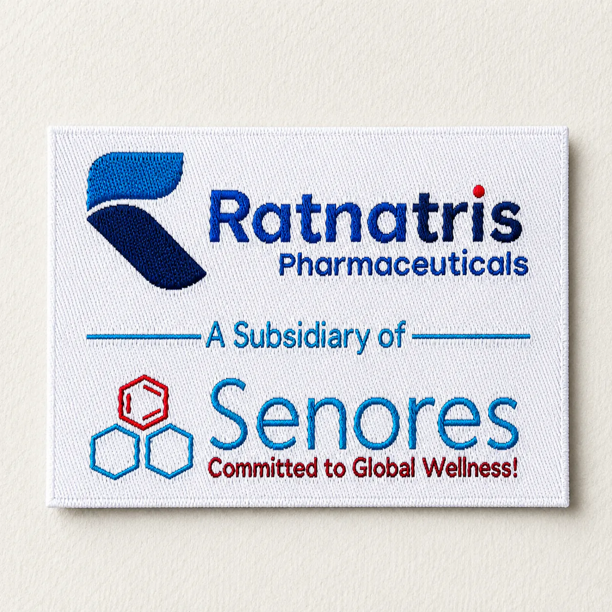 Ratnatris Pharmaceuticals white woven brand label with full-colour logo, Senores subsidiary credit and Committed to Global Wellness tagline — corporate uniform woven label sample