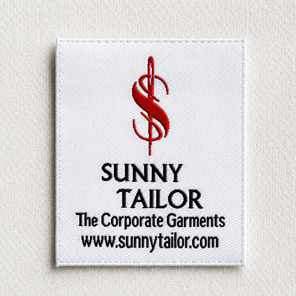 Sunny Tailor white woven brand label with red dollar-sign monogram, brand name, The Corporate Garments tagline and website URL — tailoring brand woven label sample