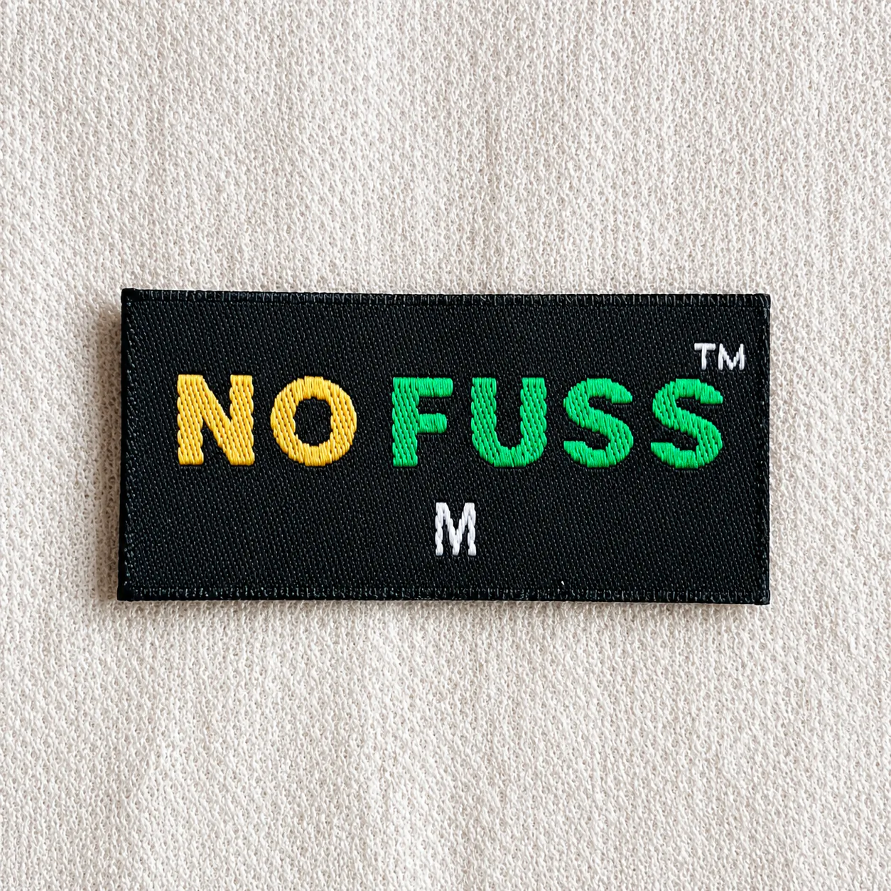 No Fuss black woven brand label with multi-colour yellow and green wordmark, TM mark and white M size marker — streetwear brand woven label sample