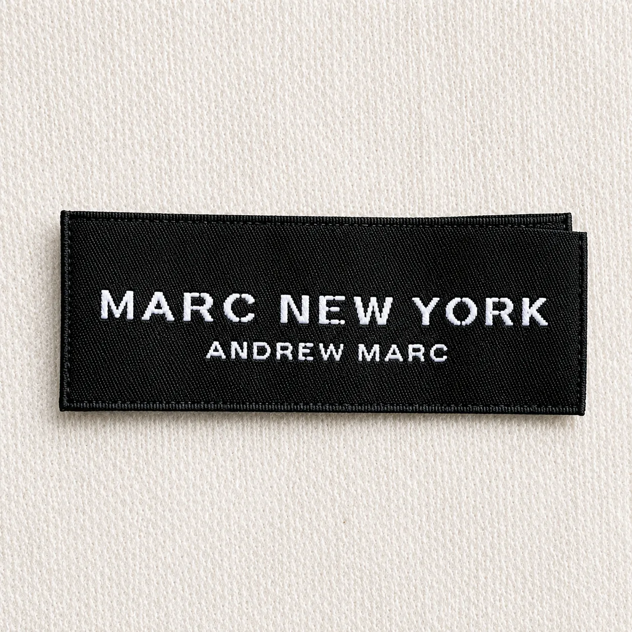 Black satin printed brand label sample with minimal white serif wordmark and secondary tagline — luxury menswear design reference, satin print finish