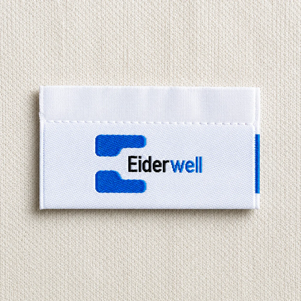 Eiderwell white centre-fold woven brand label with blue letter-E icon and italic wordmark — premium apparel woven neck label sample