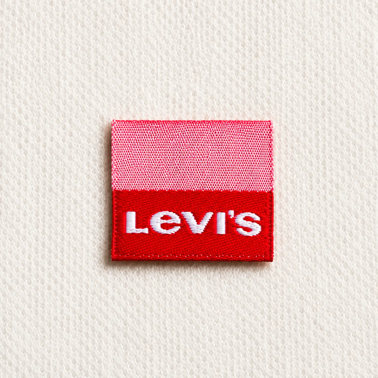 Small pink-and-red woven brand label sample with bold white block-letter wordmark — denim and casual-wear design reference, woven label finish