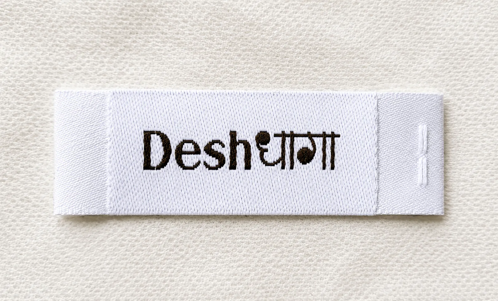 Desh white centre-fold woven brand label with English and Devanagari script wordmark — ethnic and bilingual brand woven label sample